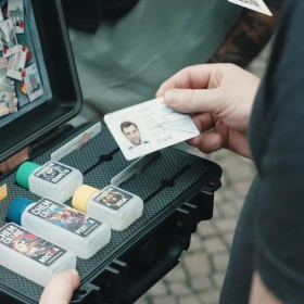 aufgeklappte Kiste mit verschiedenen Hinweismaterialien. Eine Person fällt einen Personalausweis in der Hand