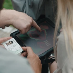Eine Frau Fällt eine schwarze aufgeklappte Kiste in der Hand. Eine andere Person hält einen Personalausweis. Eine dritte Hand zeigt auf eine Darstellung in der Kiste.