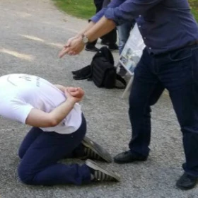eine Szene aus einem Kriminalfall wird nachgespielt. Eine Person kniet auf dem Boden mit den Händen auf dem Rücken. Eine andere Person stellt eine Pistole mit zwei Fingern dar und ziel auf die Person auf dem Boden.