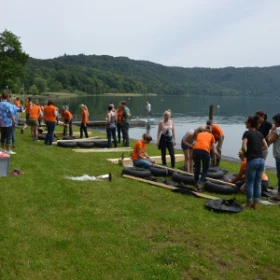 viele Personen bauen mehrere Flöße auf einer Wiese an einem See