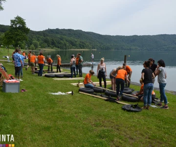 viele Personen bauen mehrere Flöße auf einer Wiese an einem See