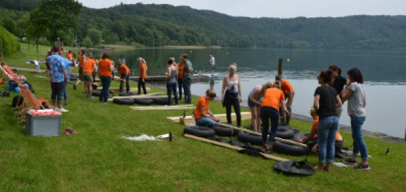 viele Personen bauen mehrere Flöße auf einer Wiese an einem See