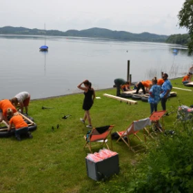 Floßbau am See, 3 Liegestühle, Personen schauen aufs Wasser