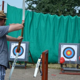 Eine Person steht mit einem Bogen in der Hand mit Blickrichtung zur Zielscheibe. Im Hintergrund ist ein grünes Fangnetz aufgebaut. Die Person hat den Pfeil gespannt und zielt auf die Zielscheibe.