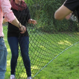 EVINTA_Teamspiel_Teamnettz_Bild 03 Drei Personen stehen um ein senkrecht gespanntes Netz herum. Eine Person steht hinter dem Netz. Das Netz hat große Löcher.