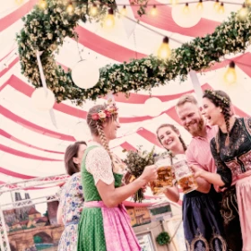 Menschen feiern Oktoberfest in einem Festzelt. Eine Gruppe steht im Kreis zusammen und stößt mit Bierkrügen an.