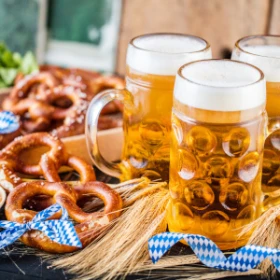 3 Bierkrüge stehen gefüllt auf einem Tisch. Daneben liegen Salzbrezeln und bayrische Dekoration.