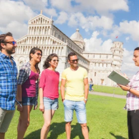 Vier Personen stehen mit einem Stadtführer auf einer Wiese vor einem weißen Gebäude. Der Stadtführer erzählt etwas und hält eine Karte in der Hand.