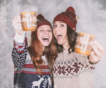 zwei Frauen mit Winterpullover und Glühweintassen in der Hand jubeln in die Kamera