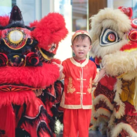Kleiner junge mit traditioneller roter chinesischer Tracht steht zwischen zwei chinesischen Figuren und feiert mit einem chinesischen Löwentanz