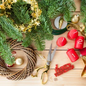 Übersicht an Materialien zur Gestaltung eines Adventskranzes, Rohling aus Holz, Tannengrün, Dekorationselemente, goldenes Band, rote Kerzen versehen mit den Zahlen 1 und 2, goldene Schneere