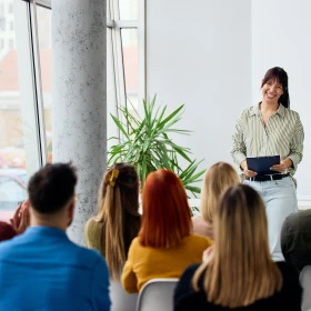 Frau steht vor einer Gruppe von Menschen und hält einen Workshop / Vortrag, Zuhörer sitzen in Reihen auf Stühlen