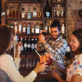 zwei Frauen stoßen mit Cocktail an der Bar an, dahinter der Barkeeper