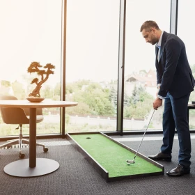 Ein Mann hat eine kleine mobile Golfbahn neben seinen Schreibtisch auf den Bogen gelegt und Spiel Golf vor einer großen Fensterfront.