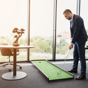 Ein Mann hat eine kleine mobile Golfbahn neben seinen Schreibtisch auf den Bogen gelegt und Spiel Golf vor einer großen Fensterfront.