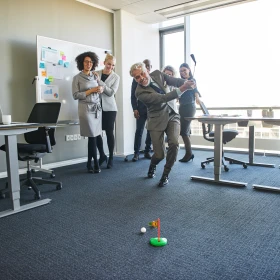 eine Gruppe Menschen spielt Golf in einem Büro. Ein Mann macht gerade einen Abschlag. Im Hintergrund jubeln Menschen.