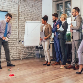 Eine Gruppe Menschen steht in einem Büro mit einer Backsteinwand. Ein Mann spiel Golf und zielt gerade auf eine kleine rote Zielflagge.