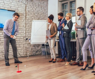 Eine Gruppe Menschen steht in einem Büro mit einer Backsteinwand. Ein Mann spiel Golf und zielt gerade auf eine kleine rote Zielflagge.