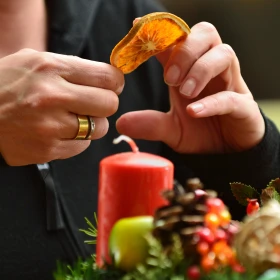 Zwei Hände halten eine Scheibe getrocknete Orange über einer roten Kerze auf einem Adventskranz