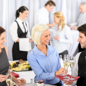 Kellner verteilt Sekt mit einen Tablett, Frau mit blauer Bluse nimmt ein Sektglas vom Tablett