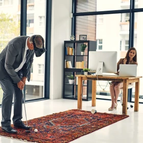 ein Mann spielt auf einem roten Teppich mit einem weißen Golfball. Der Teppich liegt in einem Büro. Der Mann trägt eine VR-Brille. Gegenüber sitzt eine Frau an einem Schreibtisch. Auf dem Schreibtisch steht ein großer Computer und ein kleiner Laptop.