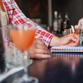 Barpersonal schreibt in ein Buch, ein Cocktail steht auf der Bar