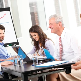 Drei Personen bei einer Besprechung vor einem Whiteboard, Auf dem Tisch ein Laptop