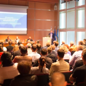 Tagung in einem großen Saal, Vortrag wird gehalten, Präsentation auf einer Leinwand