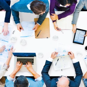 Viele Personen sitzen an einem Tisch, Tablets und Laptop sowie ausgedruckte Grafiken liegen auf dem Tisch