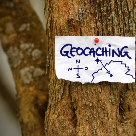 Schild mit "Geocaching" und 3 Symbolen hängt an einem Baum