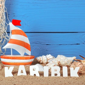 Schriftzug "Karibik" steht im Sand, Muscheln, kleines Holzboot