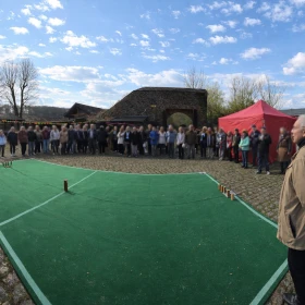 Dieses Bild zeigt eine mittelalterliche Kubb-Spielstation auf einem Burghof mit authentischer Atmosphäre und leicht aufgehelltem Wetter. Besucher stehen um das Spielfeld und beobachten das traditionelle Wurfspiel in historischer Umgebung. Die Szene vermittelt ein lebendiges Ritterturnier-Erlebnis mit Fokus auf Geschicklichkeit und Teamspaß.