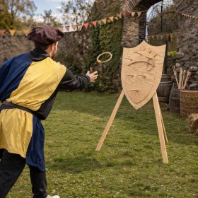 Ein mittelalterliches Ringwurf-Spiel findet auf einer grünen Wiese vor einer historischen Burg statt. Eine Person in passender Gewandung wirft gerade einen Ring auf das Ziel, was Dynamik ins Bild bringt. Die Szene verbindet Natur, Bewegung und mittelalterliche Unterhaltung.