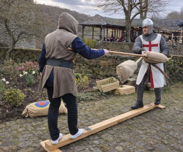 Zwei Personen treten beim mittelalterlichen Lanzenstechen auf einem Balancierbalken gegeneinander an. Die Szene zeigt Spannung, Bewegung und mittelalterlichen Wettkampfcharakter.