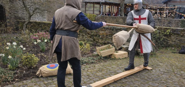 Zwei Personen treten beim mittelalterlichen Lanzenstechen auf einem Balancierbalken gegeneinander an. Die Szene zeigt Spannung, Bewegung und mittelalterlichen Wettkampfcharakter.
