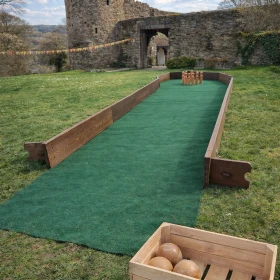 Eine klassische Kegelbahn aus Holz wurde in eine mittelalterliche Wiesenlandschaft vor einer Burg integriert. Die Szene zeigt das Spielgerät mit natürlichen Materialien und historischer Kulisse.