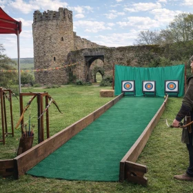 Ein mittelalterlicher Bogenschießstand mit Zielscheiben steht vor einer Burganlage in idyllischer Landschaft. Ein Teilnehmer in historischer Kleidung bereitet sich auf den Schuss vor und verleiht der Szene Lebendigkeit. Das Bild kombiniert Aktivität, Natur und authentisches Event-Feeling.