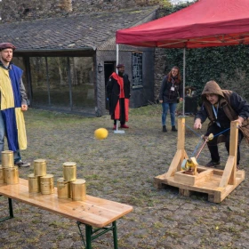 Dieses Bild zeigt eine mittelalterliche Katapult-Station, bei der ein Ball auf gestapelte Blechdosen geschossen wird. Die Szene wurde dynamisch gestaltet, sodass der Flug des Balls und die Aktion im Mittelpunkt stehen. Zuschauer in Gewandung verstärken die authentische Turnier-Atmosphäre.
