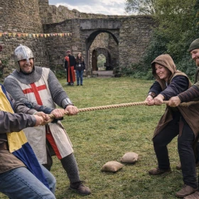 Mehrere Personen in mittelalterlicher Kleidung treten beim Tauziehen gegeneinander an und zeigen vollen Körpereinsatz. Die Szene spielt vor einer Burg und vermittelt Teamgeist, Kraft und Spaß. Ideal zur Darstellung aktiver Gruppen-Challenges auf einem Ritterturnier.