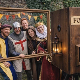 Eine Gruppe von Teilnehmern in mittelalterlichen Kostümen posiert lachend vor einer rustikalen Fotobox. Die Kombination aus Holzoptik, Requisiten und historischer Umgebung schafft eine authentische Event-Atmosphäre. Dieses Bild eignet sich perfekt für Erinnerungsfotos und interaktive Event-Highlights.