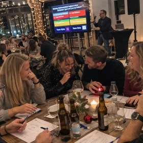 Authentische Momentaufnahme einer Quizshow-Weihnachtsfeier: Eine Gruppe von Kollegen sitzt eng beisammen an einem Tisch, lacht ausgelassen und diskutiert die nächste Antwort. In der Mitte liegt ein Buzzer, während im Hintergrund ein Moderator vor einem Bildschirm mit Quizfragen steht. Die warme Beleuchtung, Weihnachtsdeko und gefüllten Gläser vermitteln eine entspannte, gesellige Atmosphäre wie bei einem echten Event vor Ort.