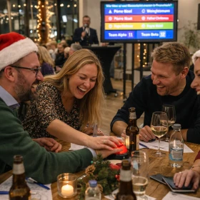 Lebendige Szene aus einer laufenden Quizrunde: Vier Teilnehmer beugen sich gemeinsam über den Tisch, während eine Person gerade den Buzzer drückt. Die Gruppe reagiert spontan mit Lachen und Begeisterung. Getränke, Notizen und kleine weihnachtliche Details auf dem Tisch unterstreichen den authentischen Charakter der Veranstaltung, während im Hintergrund weitere Gäste und der Quizbildschirm zu sehen sind.