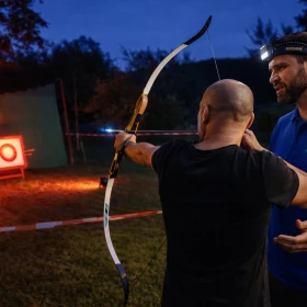 Das Bild zeigt einen Teilnehmer beim LED-Nachtbogenschießen auf einem Firmengelände, der konzentriert auf beleuchtete Zielscheiben schießen. Hinter den Scheiben sorgt ein durchgehendes Fangnetz für Sicherheit, während die LED-Beleuchtung der Ziele und Pfeile eine eindrucksvolle Lichtstimmung erzeugt. Die Szene vermittelt eine professionelle, moderne Eventatmosphäre und unterstreicht den Teambuilding-Charakter dieses besonderen Firmenevents.
