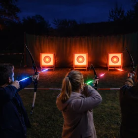 Das Bild zeigt ein stimmungsvolles LED-Nachtbogenschießen auf einem Firmengelände in den Abendstunden. Drei Teilnehmer stehen nebeneinander auf einer markierten Schusslinie und richten konzentriert ihre Bögen auf die hell beleuchteten Zielscheiben in einiger Entfernung. Die Pfeile leuchten in unterschiedlichen LED-Farben – Blau, Grün und Rot – und erzeugen eine eindrucksvolle Lichtdynamik in der Dunkelheit.