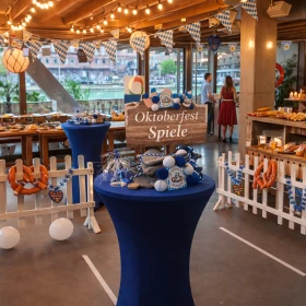 Die Aufnahme präsentiert eine stilvoll dekorierte Indoor-Location im Oktoberfest-Stil mit Buffet, Spielstationen und bayerischer Atmosphäre als hochwertiges Firmenevent im B2B-Bereich. Das Setting unterstreicht die Kombination aus Teambuilding, Oktoberfest-Spielen und kulinarischem Rahmenprogramm und eignet sich ideal für Mitarbeiterveranstaltungen, Incentives oder Firmenfeiern bundesweit.