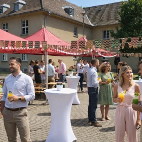 Unter roten Zelten mit bunten Wimpelketten genießen Gäste erfrischende Getränke und tauschen sich an Stehtischen aus. Die dekorierten Flächen und das helle Tageslicht schaffen eine fröhliche Sommerfest-Stimmung.