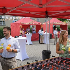 Unter einem roten Zeltdach stehen Gäste beisammen, unterhalten sich und genießen bunte Drinks, während im Hintergrund weitere Stände aufgebaut sind. Die lebendige Szenerie vermittelt eine lockere, gemeinschaftliche Atmosphäre.