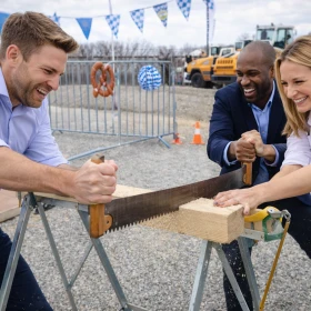 Zwei Teammitglieder sägen gemeinsam durch einen Holzbalken und geben dabei alles, um als Erste das Ziel zu erreichen. Lachende Kolleginnen und Kollegen begleiten das Kräftemessen mit Begeisterung.